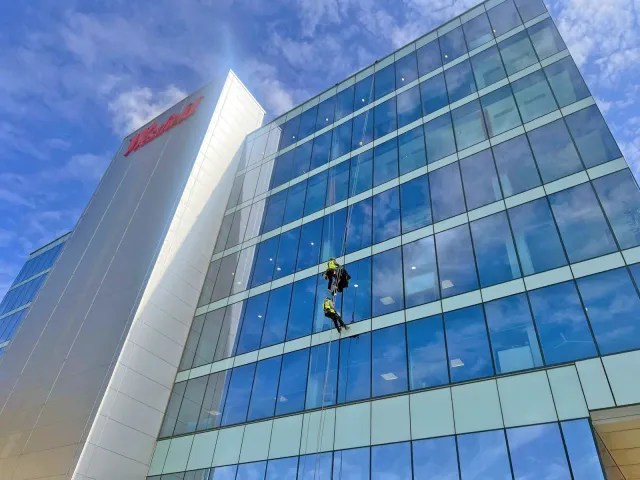 Two rope access technicians carrying out high-level glazing work on the glass façade of a modern commercial building.
