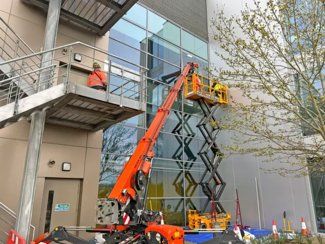 Commercial Window Repair Using Mobile Access Equipment Technicians using a mobile crane and scissor lift to carry out commercial window repair work on a modern glass-fronted building.