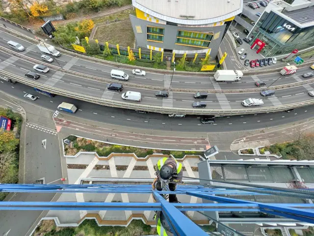 Rope Access Safety Work Above Busy Road Rope access technician working at height on a blue building frame above a busy road and commercial area.