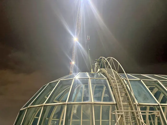 Night-Time High-Level Glazing Work on Glass Atrium Roof Rope access technician working at night on a glass atrium roof with crane lighting overhead during high-level glazing maintenance.