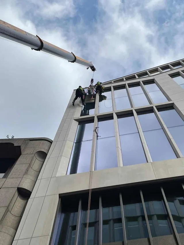 High-Level Glass Replacement Using Rope Access and Crane Support Rope access technicians replacing a high-level glass panel on a modern commercial building with support from a mobile crane.