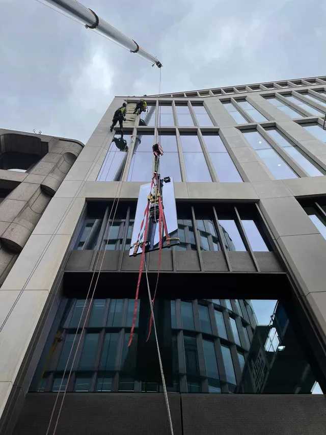 Rope Access Glass Replacement on Commercial Building Rope access technicians using crane support to lift and position a replacement glass panel on the upper floors of a modern commercial building.