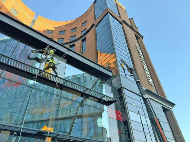Rope access technicians inspecting and working on high-level glazing at a commercial building, with a mobile access platform supporting work on the upper façade.